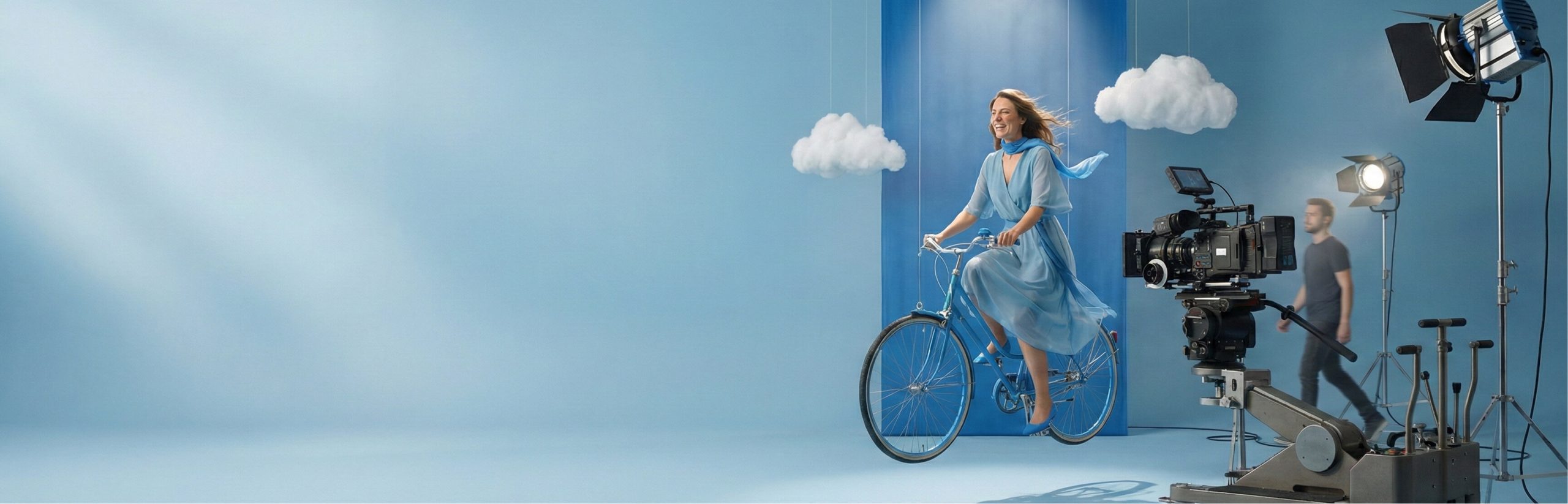 A woman in a flowing blue dress rides a bicycle on a pale blue film set with paper clouds, as a camera crew films her